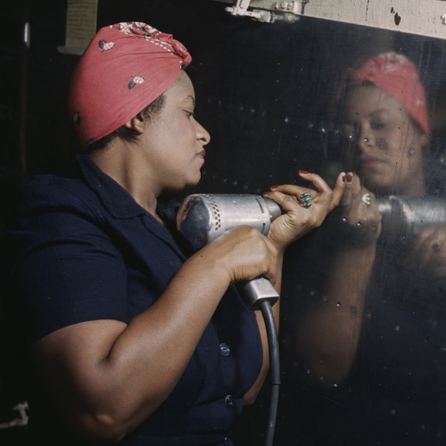 Photograph of African American woman in red headscarf and blue coveralls working on a “Vengeance” dive bomber with a hand drill. 1943